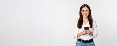 Young woman with smartphone, smiling and looking at camera, using mobile phone app, cellular technology and online shopping concept, white background.