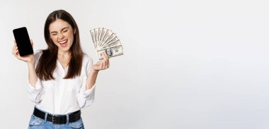 Enthusiastic young woman winning money, showing smartphone app interface and cash, microcredit, prize concept, standing over white background.