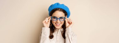 Close up portrait of stylish woman posing in sunglasses, laughing and smiling, wearing trendy outfit, standing over beige background.