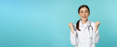 Happy and excited young female doctor, physician celebrating, achieve goal and smiling pleased, winning, standing in white coat against blue background.