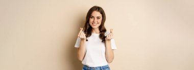 Cute young woman showing finger hearts sign and smiling, express care and love, standing over beige background.