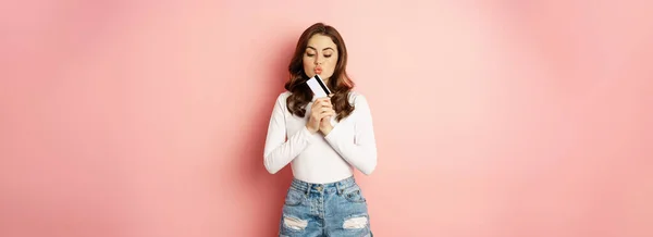 スタイリッシュな若い女性は彼女のクレジットカードにキスをし 買い物をするのが好きで ピンクの背景に立っています スペースのコピー — ストック写真