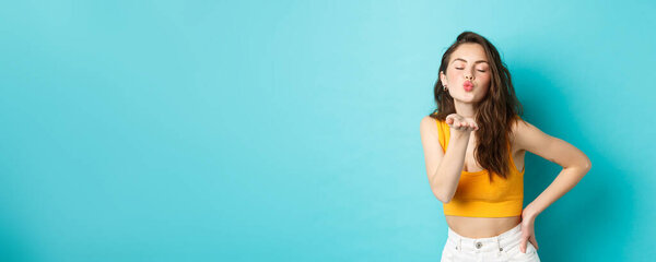 Coquettish summer girl sending air kiss at camera, pucker lips and close eyes, standing over blue background.