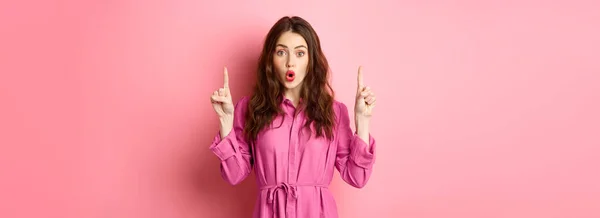 stock image Surprised young brunette woman in her 20s, saying wow, staring impressed at camera while pointing fingers up at logo, showing promo offer, standing over pink background.