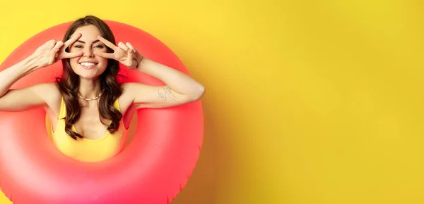 stock image Portrait of attractive modern girl inside trendy pink beach swimming ring, showing peace v-sign and smiling happy, standing over yellow background.