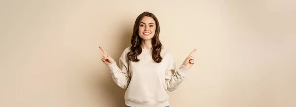 stock image Cheerful caucasian woman showing directions, two ways, pointing sideways at variants, choices for customer, standing over beige background.