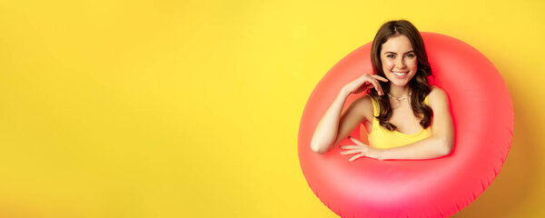 Close up portrait of beautiful brunette woman model inside pink swimming ring, beach accessories and summer vacation concept, yellow background.