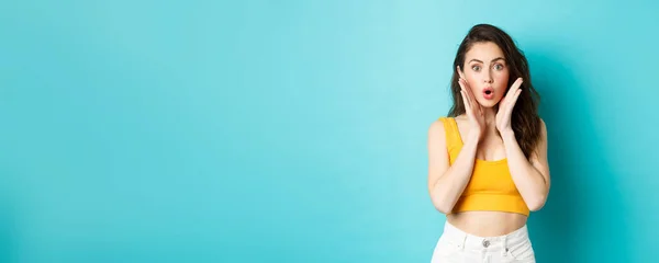 stock image Summer and lifestyle concept. Portrait of brunette girl looks surprised, gasping and holding hands near face, staring in awe at camera, checking out big news, blue background.