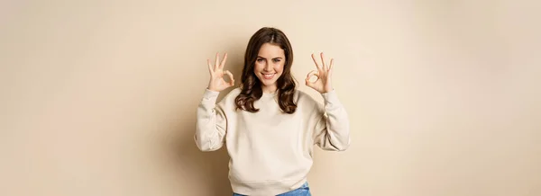 stock image Okay. Confident smiling modern girl showing ok sign, approve, like and recommend smth good, standing over beige background.