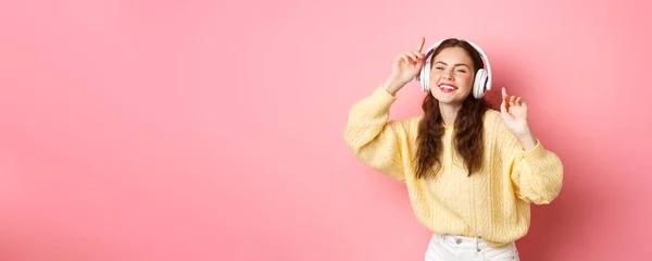 stock image Beautiful stylish female model singing favorite song, listening to music in wireless headphones, smiling and dancing, standing over pink background.