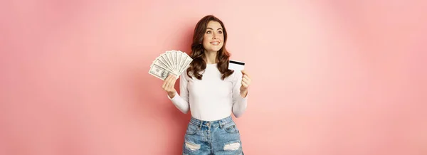 お金および貸付け金の概念 幸せな美しい女の子を保持クレジットカードと現金 満足そうな ショッピングを考えて ピンクの背景の上に立って — ストック写真