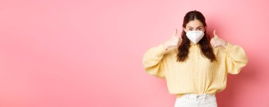 Covid-19, tecrit ve salgın konsepti. Solunum cihazı takan genç bir kadın, karantina sırasında yüz maskesi takan, onay verirken baş parmağını gösteren, aşıyı destekleyen, pembe arka plan.