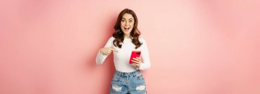 Happy brunette girl showing announcement, pointing finger at mobile phone, showing online promo, app news, standing against pink background.