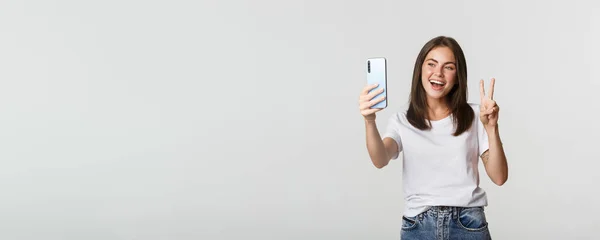 Atrakcyjna Brunetka Wygląda Szczęśliwie Robi Selfie Smartfonie Pokazując Gest Pokoju — Zdjęcie stockowe