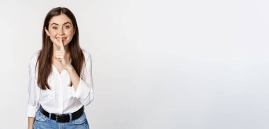 Smiling beautiful woman shushing, making hush shh sign, press finger to lips and winking, hiding secret, standing in blouse and jeans over white background.