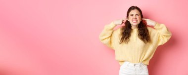 Sinirli genç bir kadın üstteki komşulara küfrediyor, yukarı bakıyor, kulakları kulaklarıyla tıkanmış, gürültüden şikayetçi, müzikten şikayetçi, pembe arka planda duruyor..