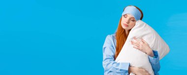Güzellik, kadın ve yaşam tarzı konsepti. Rahat ve rahatlamış uyku maskesi ve pijama giymiş güzel kızıl saçlı kadın, uykulu gözler, tatlı rüyalar görüyor, yastığa sarılıyor, mavi arka planda dikiliyor..