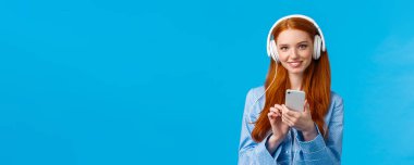 Nazik ve kadınsı, sevimli kızıl saçlı kız öğrenci, kulaklık takıyor klasik müzik okuyor, mavi arka planda duruyor, gece elbisesi giyiyor. Boşluğu kopyala