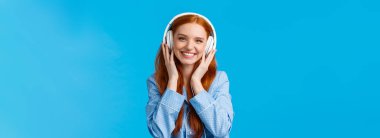 Belden yukarısında neşeli seksi beyaz kız büyük kulaklıklarla müzik dinliyor, gülümsüyor, arkadaşlarına bakıyor, bekâr, gece kıyafeti giyiyor, mavi arka planda duruyor..