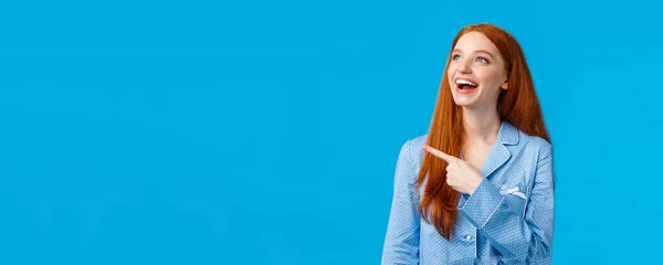 stock image Delighted and entertained good-looking caucasian redhead girl in nightwear, wearing pyjama to sleep, pointing looking upper left corner amused, laughing happily, standing blue background.