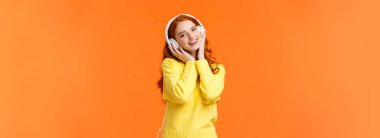 Bel yukarı portresi, beyaz kulaklıklı tatlı kızıl saçlı kız, eğik kafalı müzik, kulaklıklar ve gülümseyen kamera, en sevdiği şarkı boyunca şarkı söyleyen, turuncu arka planda duran. Boşluğu kopyala