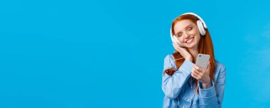 Şefkat, kadınlar ve yaşam tarzı konsepti. Pijama giymiş neşeli kızıl saçlı kadın, başını eğmiş müzik dinlemekten zevk alıyor, akıllı telefon tutuyor ve kulaklık gibi gülümsüyor..
