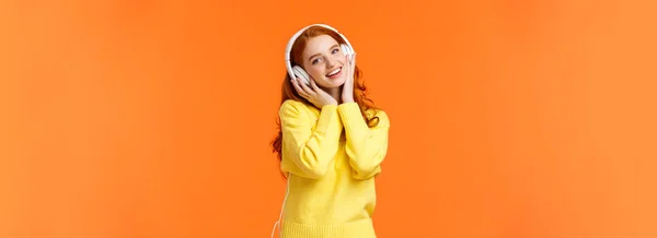 穿着白色耳机的可爱的红头发少女的蜡像 倾斜的头听音乐 触摸耳机和微笑的相机 唱着最喜欢的歌曲 站在橙色的背景上 复制空间 — 图库照片