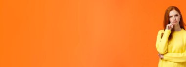 Şüpheci ve kararsız kızıl saçlı, ciddi görünümlü kadın zor kararlar alıyor, seçim hediyesi veriyor, kış tatilini geçiriyor, alışveriş koridorlarına bakıyor, dudaklarına dokunuyor, düşünüyor, turuncu arka plan düşünüyor..