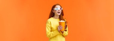 Patlamış mısır yiyen, gülümseyen, büyük ekranda film seyreden, ağzı açık, heyecanlı, fantezi sinemasında 3D gözlük takan, turuncu arka planda duran bir kız..