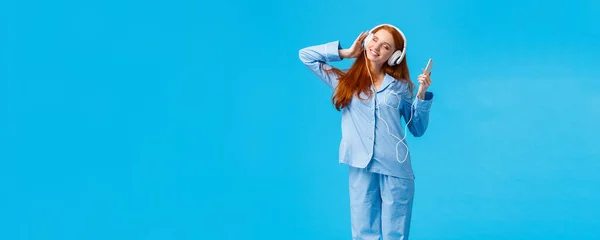 多么美丽的早晨听最喜欢的演奏会 垂直的全长肖像柔嫩的红头发女人 穿着睡衣 手持智能手机 倾斜的头和紧闭的眼睛 欣赏音乐 蓝色背景 — 图库照片