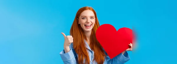 正と興奮陽気な若い赤毛の女性の親指を示す または承認 バレンタインデーには ハートカードの愛と称賛を表現し 笑みを浮かべて青の背景に大成功した — ストック写真