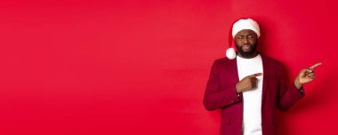 Noel, parti ve tatil konsepti. Üzgün Afro-Amerikan adam kasvetli görünüyor, gözleri kapalı ve iç çekiyor, parmaklarını sağa gösteriyor, kırmızı arka planda Noel Baba şapkasının içinde duruyor..