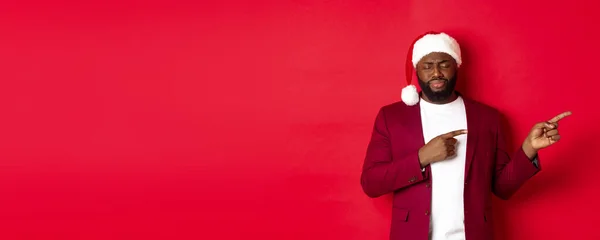 Stock image Christmas, party and holidays concept. Upset african american man looking gloomy, close eyes and sighing, pointing fingers right, standing in santa hat against red background.