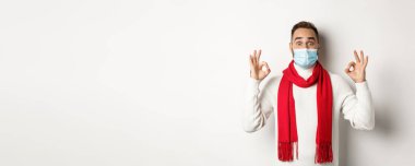 Covid-19, tecrit ve karantina konsepti. Yüz maskeli etkileyici bir adam, tamam işaretleri gösteriyor ve katılıyor, harika bir ürün öneriyor, beyaz arka plan..