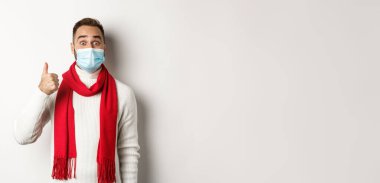 Covid-19, tecrit ve karantina konsepti. Tıbbi maskeli, şaşırtıcı ve tatmin olmuş bir adam başparmağını kaldırmış, heyecanlı görünüyor, iyi, beyaz bir arka plan gibi..