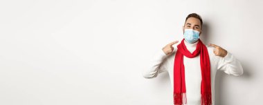 Covid-19, tecrit ve karantina konsepti. Gülümseyen adam yüz maskesi takmayı öneriyor, onu işaret ediyor, beyaz arka plana karşı duruyor..