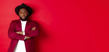 Gönülsüz ve üzgün bir siyah adam özür bekliyor, göğsünde çapraz kollar var ve kameraya kırgın bakıyor, kırmızı arka plan. Boşluğu kopyala