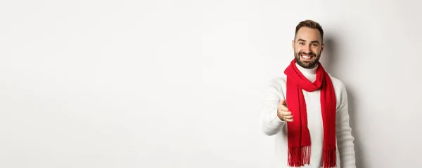 stock image Friendly young man greeting person with handshake, smiling and wishing merry christmas, wearing red scarf and winter sweater, white background.