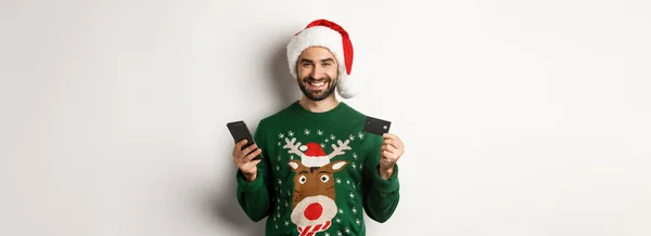 网上购物和圣诞节的概念 快乐的大胡子男人 带着信用卡和电话在网上购物 戴着圣诞帽 身穿毛衣 背景洁白 — 图库照片