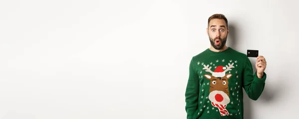 クリスマス休暇とショッピングのコンセプト 驚くべきハンサムな男でセーターに示すクレジットカード 白い背景の上に立って — ストック写真