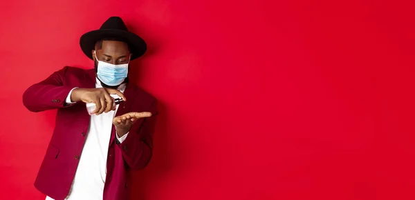 stock image Covid-19, quarantine and holidays concept. Handsome Black man in face mask and party outfit, disinfecting hands with hand sanitizer, using antiseptic, standing over red background.