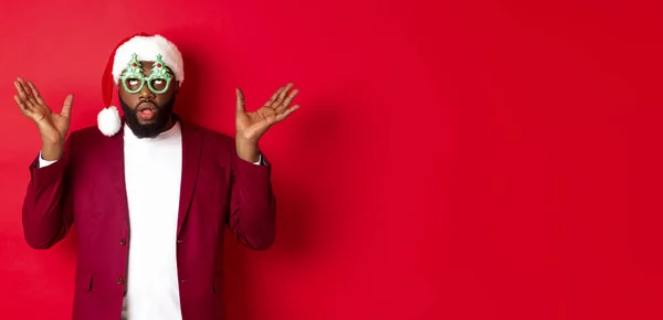 Merry Christmas. Cheerful Black man wearing funny party glasses and santa hat, smiling joyful, celebrating winter holidays, standing over red background.
