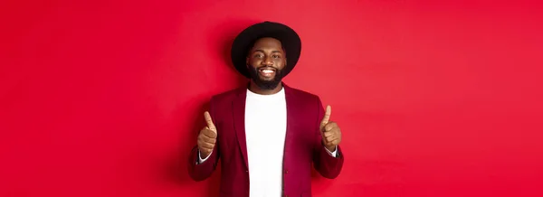 Stock image Christmas shopping and people concept. Handsome Black man smiling satisfied, showing thumbs-up, like and agree, approve something, standing against red background.