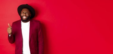 Noel alışverişi ve insanların konsepti. Yakışıklı, sakallı, parti ceketli siyahi adam başparmağını gösteriyor, gülüyor ve eğleniyor, kırmızı arka planda duruyor..