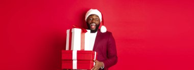 Noel, yeni yıl ve alışveriş konsepti. Neşeli siyah adam gizli noel baba hediyeler tutuyor ve gülümsüyor. Heyecanlı, hediyeler getiriyor, kırmızı arka planda duruyor..