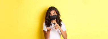 Covid-19 kavramı, sosyal mesafeler ve yaşam tarzı. El dezenfektanı kullanan, sarı arka planda duran yüzü maskeli Afro-Amerikan bir kız resmi.