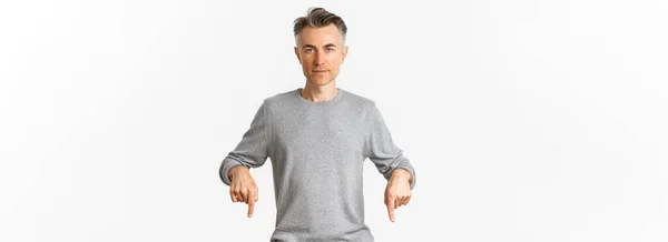 成功した中年男性の肖像画グレーのセーター 指を下に指して カメラを見て自信を持って ロゴを示す 白い背景の上に立って — ストック写真
