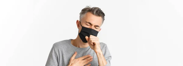Covid 社会的距離および隔離の概念 白い背景の上に立って 悪い感じ 咳や黒の医療マスクで病気の中年の男のクローズアップ — ストック写真