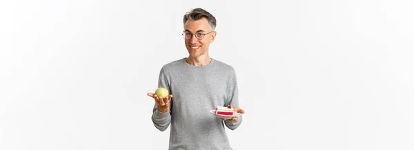 白の背景に立って ケーキの代わりにリンゴを食べることをお勧めし メガネでハンサムで健康的な中年の男性のイメージ — ストック写真