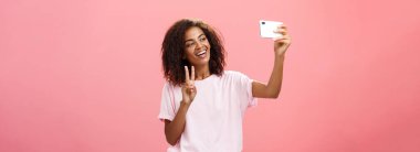 Havalı, sosyal, yakışıklı, esmer, kıvırcık saçlı kız öğrenci. Yüzüne yakın akıllı bir telefonla el ele tutuşuyor. Cihaz ekranına barış işareti yerleştirirken tasasız gülümsüyor.